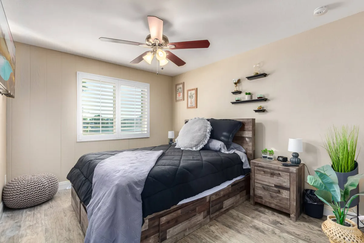 A cozy bedroom featuring a bed with a quilted cover, a window with blinds letting in soft natural light, wall shelves with Southwest-inspired decor, a nightstand with a lamp, and a potted plant beside the bed. The space feels welcoming and restful, ideal for relaxation or quiet moments.
