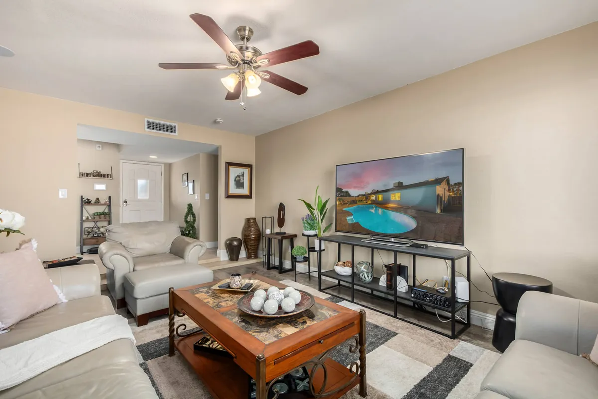 A bright, open-plan living room featuring a comfortable sofa, a coffee table, a large television, and a potted plant. Natural light pours in from windows, creating a cozy and welcoming atmosphere. The space is styled with modern Southwestern accents and is clearly designed for relaxing, entertaining guests, or watching TV together.