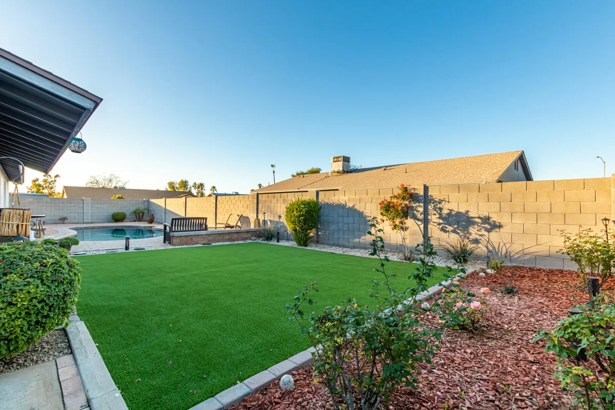A bright daytime view of a green lawn leading to a sparkling pool and covered patio. The landscaping is lush and thoughtfully arranged, with flower beds and outdoor dining areas inviting morning coffee, games, or alfresco meals.