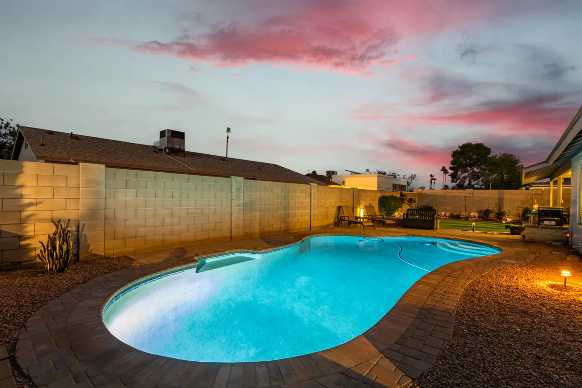 An inviting backyard scene at twilight featuring a sparkling swimming pool, an outdoor seating area, beautifully landscaped garden, and a vibrant sky. The area is softly lit, suggesting a tranquil, private atmosphere perfect for evening swims or gatherings.