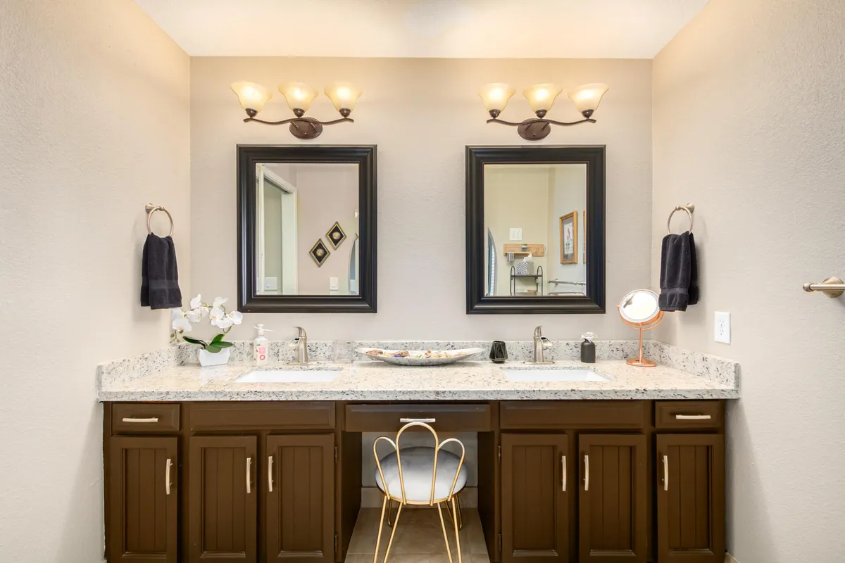 A bathroom with a double vanity, stylish mirrors, elegant lighting fixtures, a decorative bowl, and a chair tucked neatly underneath. The space is calm, organized, and ideal for shared routines or individual moments of self-care.