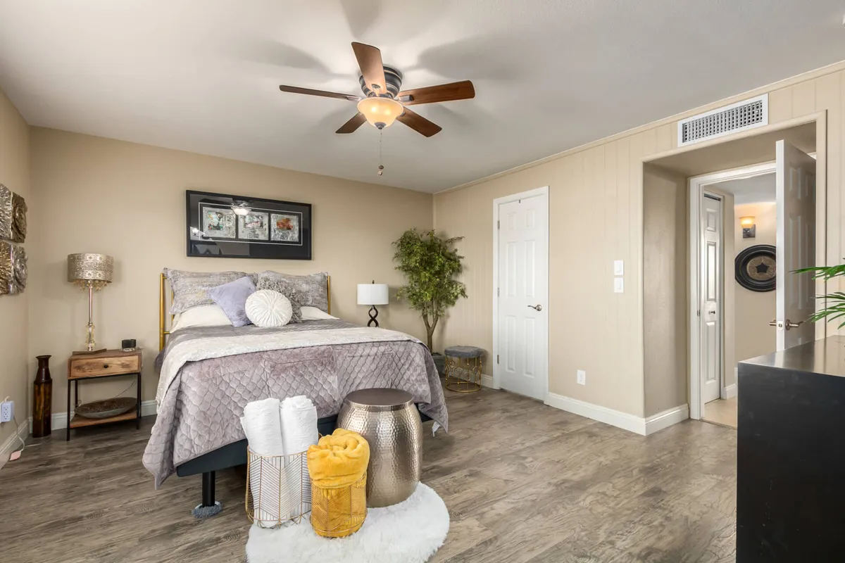 A tastefully decorated bedroom with a bed adorned with decorative throw pillows, a nightstand and lamp, wall art, an indoor plant, and an open door hinting at spaciousness. Sunlight streams in, creating a bright and peaceful atmosphere.