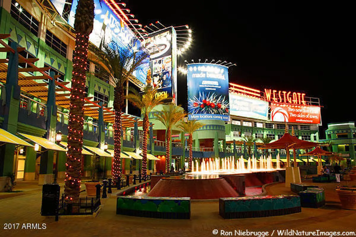 A lively nighttime scene featuring illuminated fountains with dancing water jets, vibrant billboards, palm trees wrapped in lights, and sleek modern buildings bustling with people enjoying outdoor dining and entertainment.