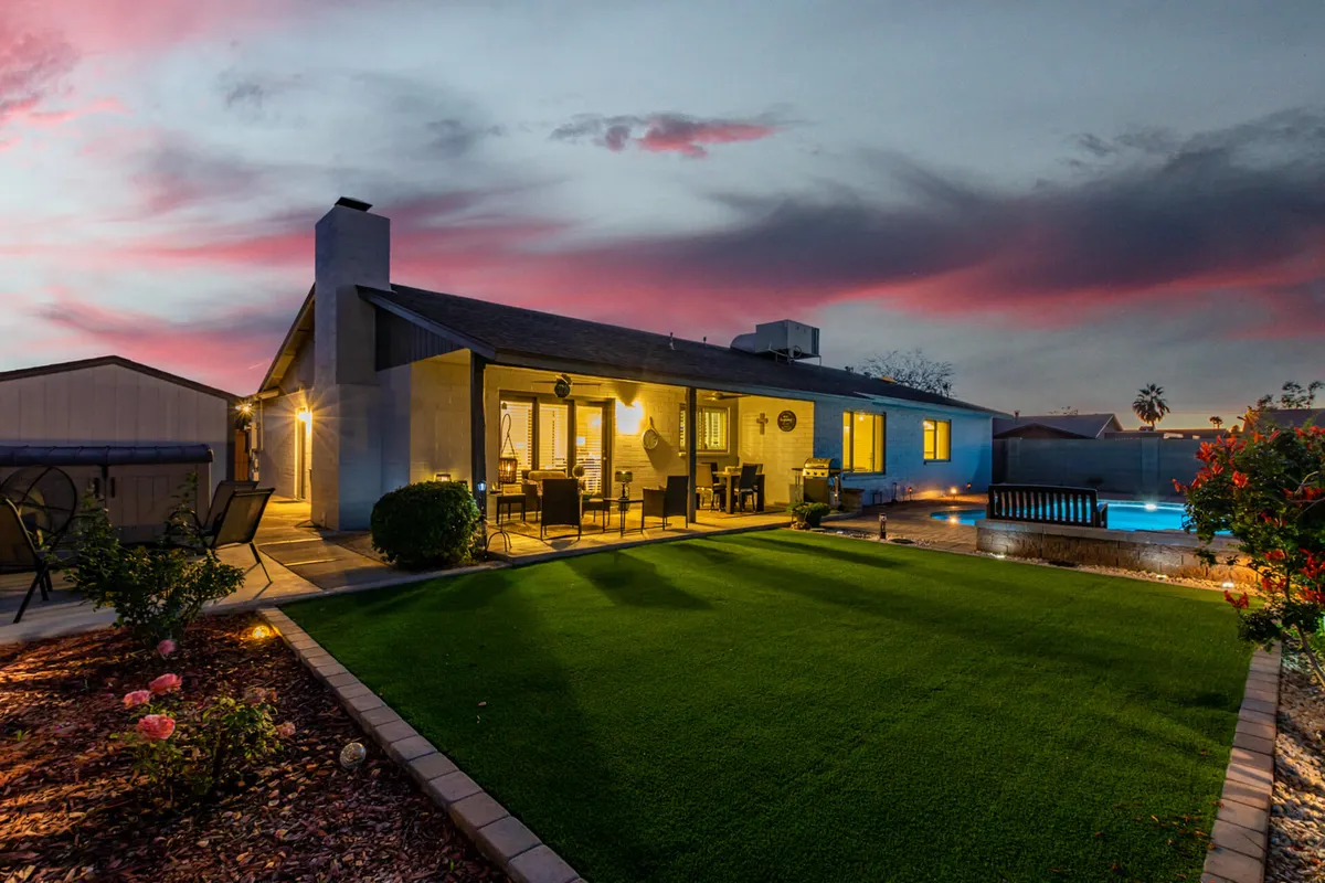 A welcoming backyard seating area overlooking a serene pool, set against the backdrop of a glowing sunset sky. Vibrant flowers and lush greenery add to the tranquil environment, while the setup is ideal for both relaxing and social gatherings.