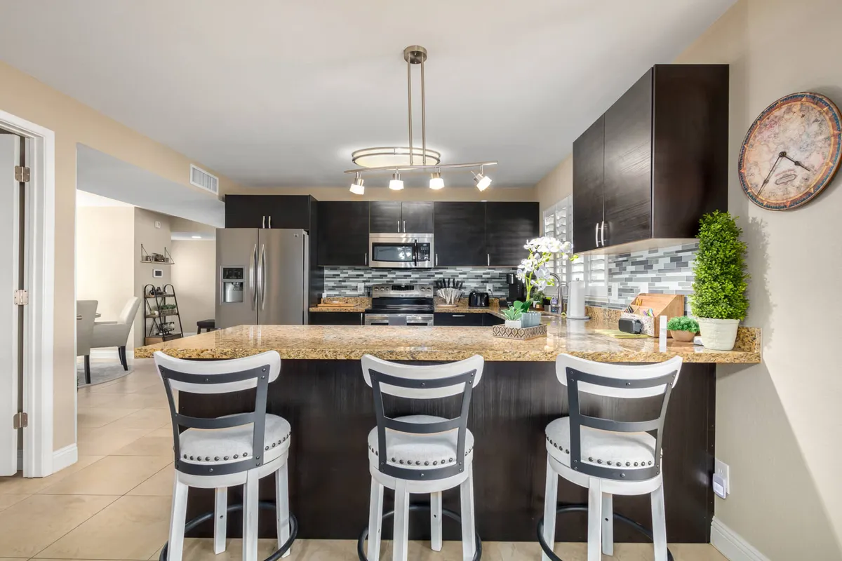 A welcoming kitchen scene with a large granite countertop, kitchen island seating, stainless steel appliances, and decorative plants. The setup suggests space for both cooking and casual dining, perfect for entertaining or enjoying a family breakfast.