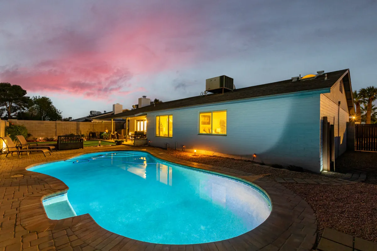 The backyard scene highlights a sparkling swimming pool, ample seating, the inviting house exterior, and a vivid sunset sky. The relaxed atmosphere suggests a perfect space to unwind after a day at games or events.