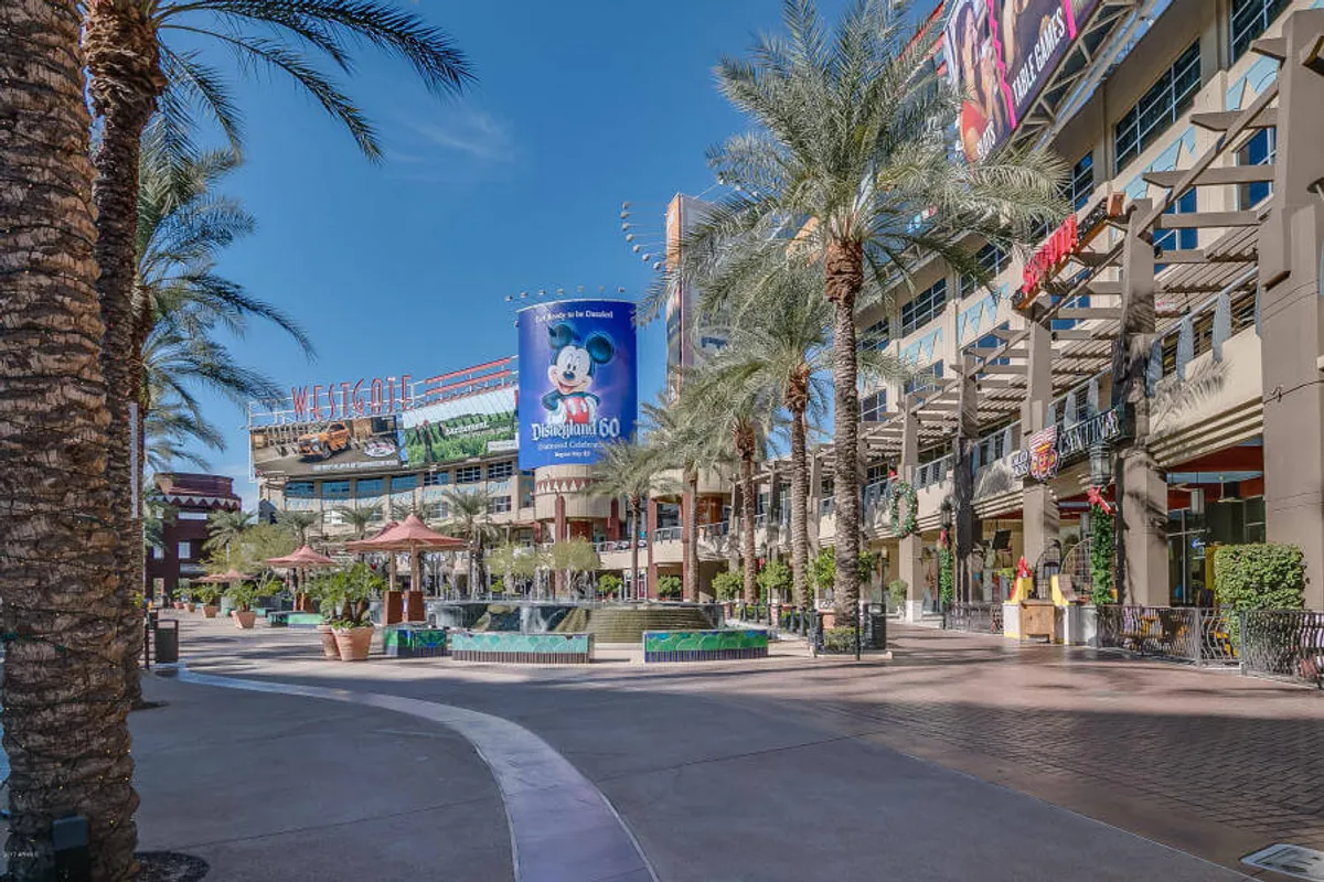 This image captures the lively Westgate Entertainment District featuring a prominent fountain, palm trees, a large billboard, and inviting shops. Shoppers and diners are visible enjoying the energetic atmosphere, hinting at the district's role as a local hub for shopping, dining, and entertainment.