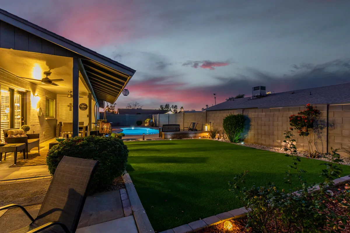 A tranquil backyard scene showcasing a sparkling private pool surrounded by lush landscaping and mature palm trees. The covered patio features comfortable seating, and the entire space is bathed in the warm glow of a colorful sunset, creating an inviting environment for evening gatherings or peaceful relaxation.